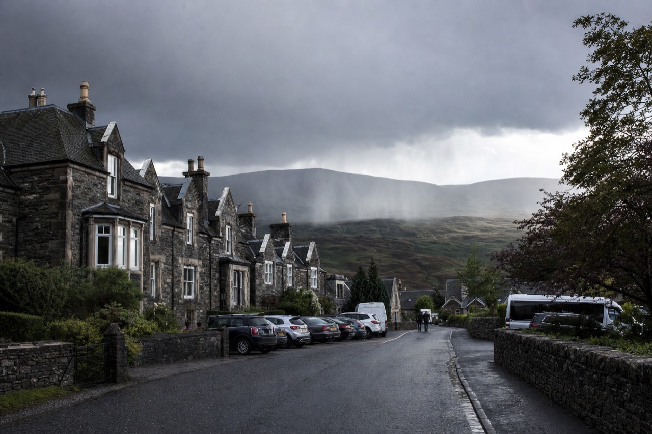 A stone Highland village street with a small hotel and parked cars under grey afternoon light.