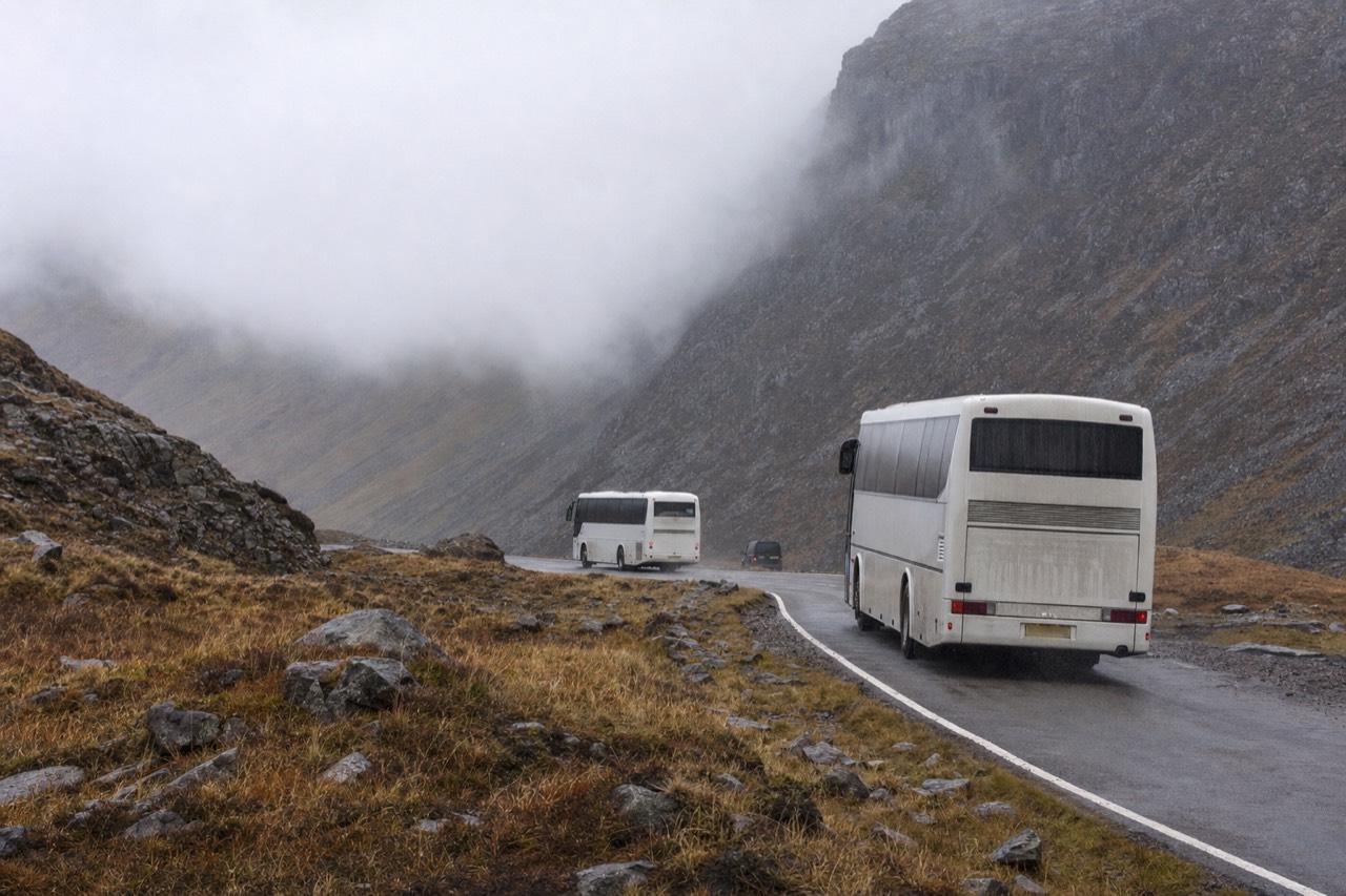 A coach on a Highland pass road with steep scree slopes rising on either side and cloud sitting on the ridge.